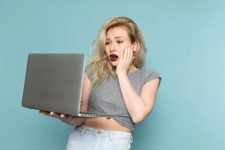 A young businesswoman looks distressed while using a slow Windows laptop. She's wearing a gray top and has blonde hair, set against a teal background. Her hand is on her face, expressing frustration as she struggles to complete an important task due to computer performance issues.